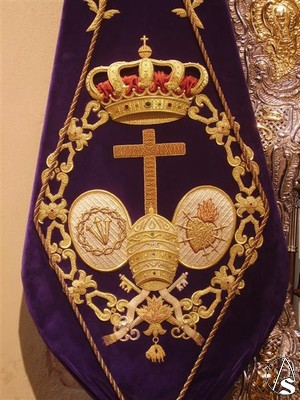  El escudo de la hermandad lo forma la cruz de animas y a sus lados un ovalo, en uno de ellos la corona de espinas y los tres clavos y en el otro un corazn con los siete puales, rodea todo ello el toisn de oro y remata la corona real. 